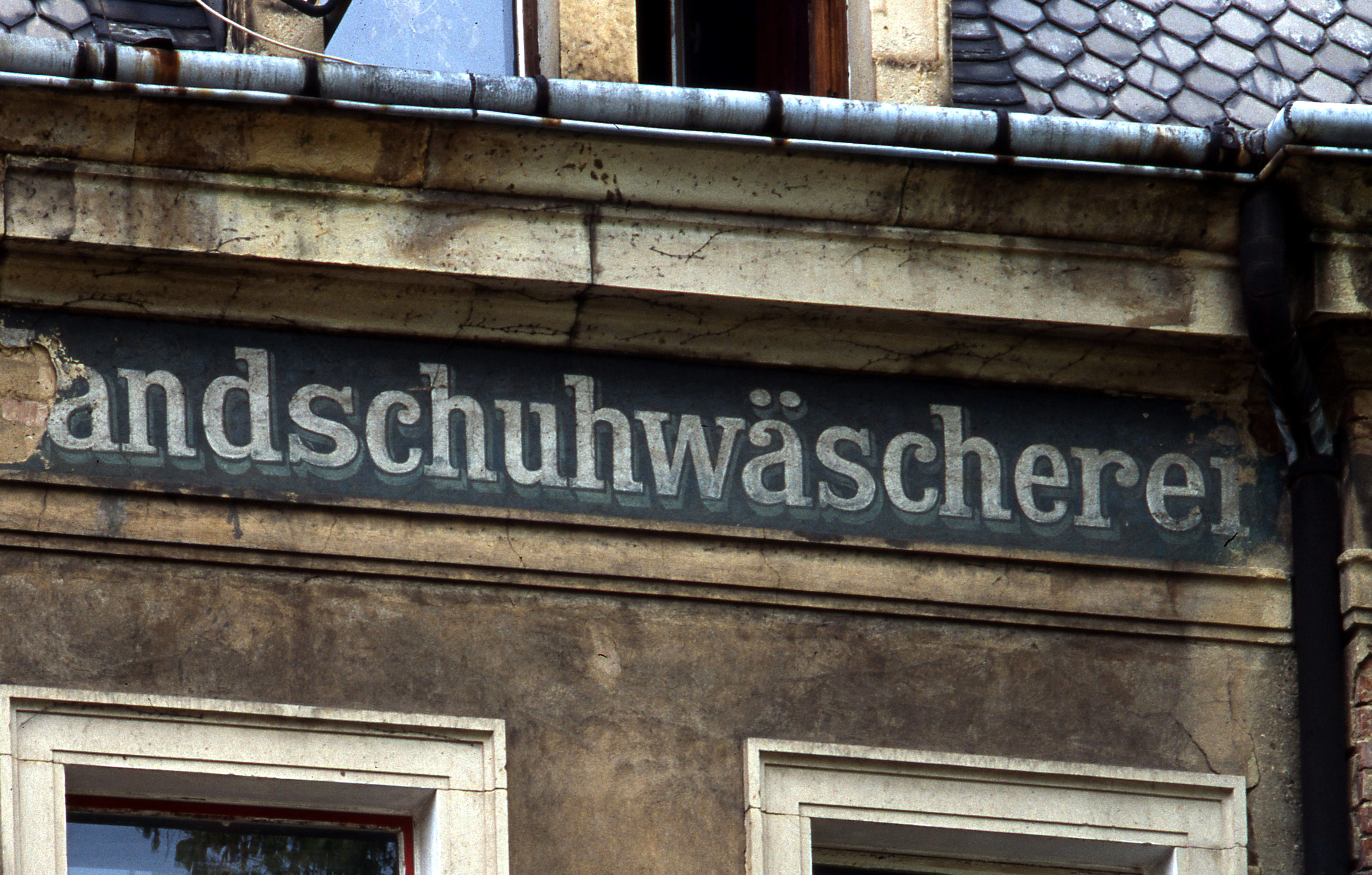 Ansicht einer verfallenen Fassade. Unter dem Dach ist der verwitterte Schriftzug "Handschuhwäscherei" zu erkennen.