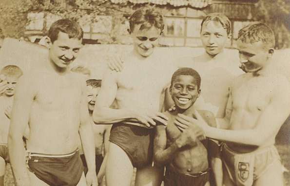 4 Jugendliche stehen 1914 im König-Friedrich-August-Bad in Klotzsche und lachen in die Kamera. In ihrer Mitte steht ein Junge mit dunkler Hautfarbe, der ebenfalls lacht. Zwei der Jugendlichen fassen den Jungen am Oberkörper an, vermutlich sind sie fasziniert von seiner Hautfarbe.