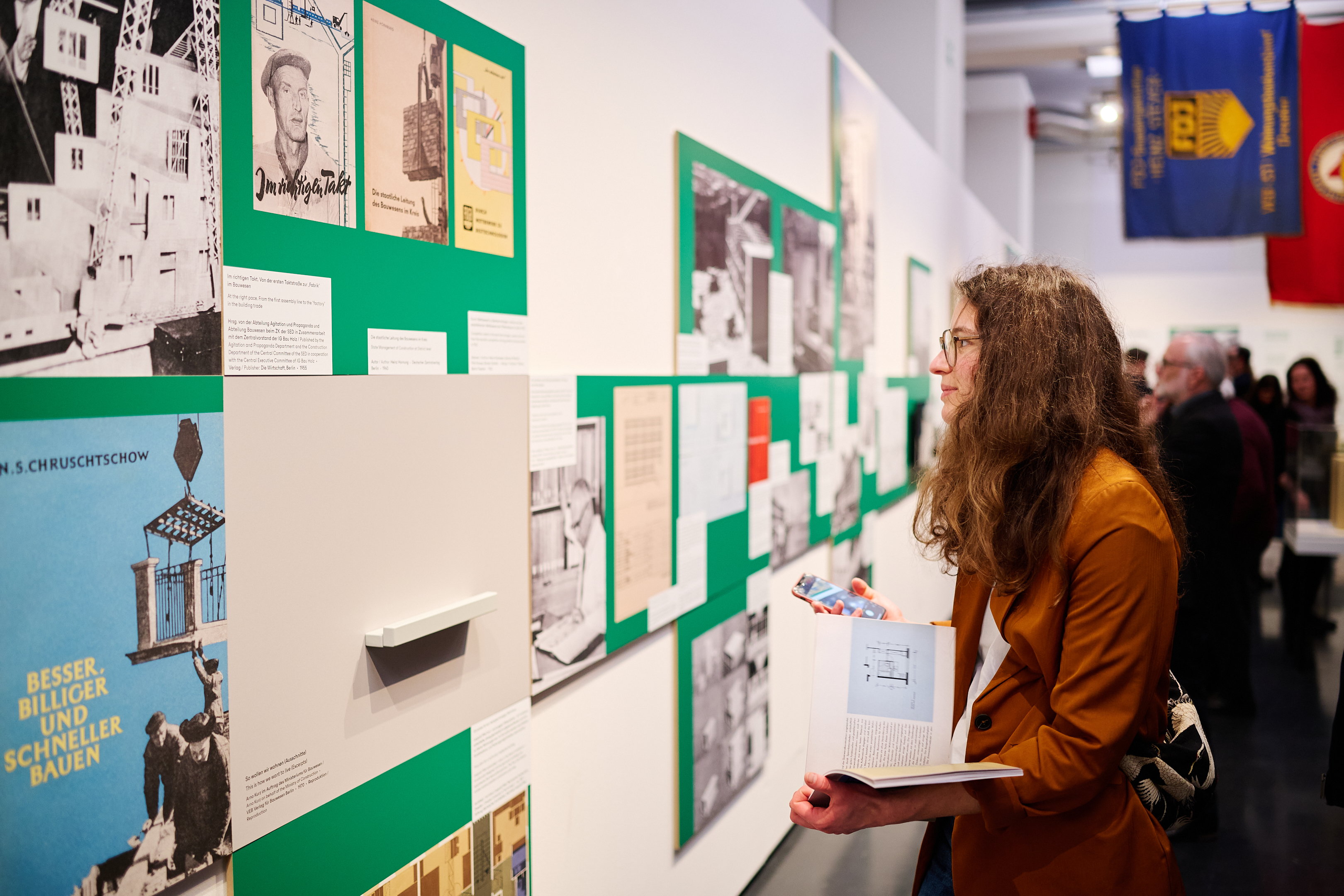 KI generiert: Das Bild zeigt eine Person, die in einer Ausstellung aufmerksam Wandtafeln mit historischen Fotografien und Dokumenten betrachtet. Im Hintergrund sind weitere Besucher und farbige Banner sichtbar.