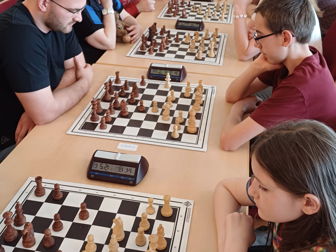 KI generiert: Das Bild zeigt mehrere Personen, die Schach an Tischen spielen. Jeder Tisch hat ein Schachbrett und eine Schachuhr, während die Spieler konzentriert nachdenken.