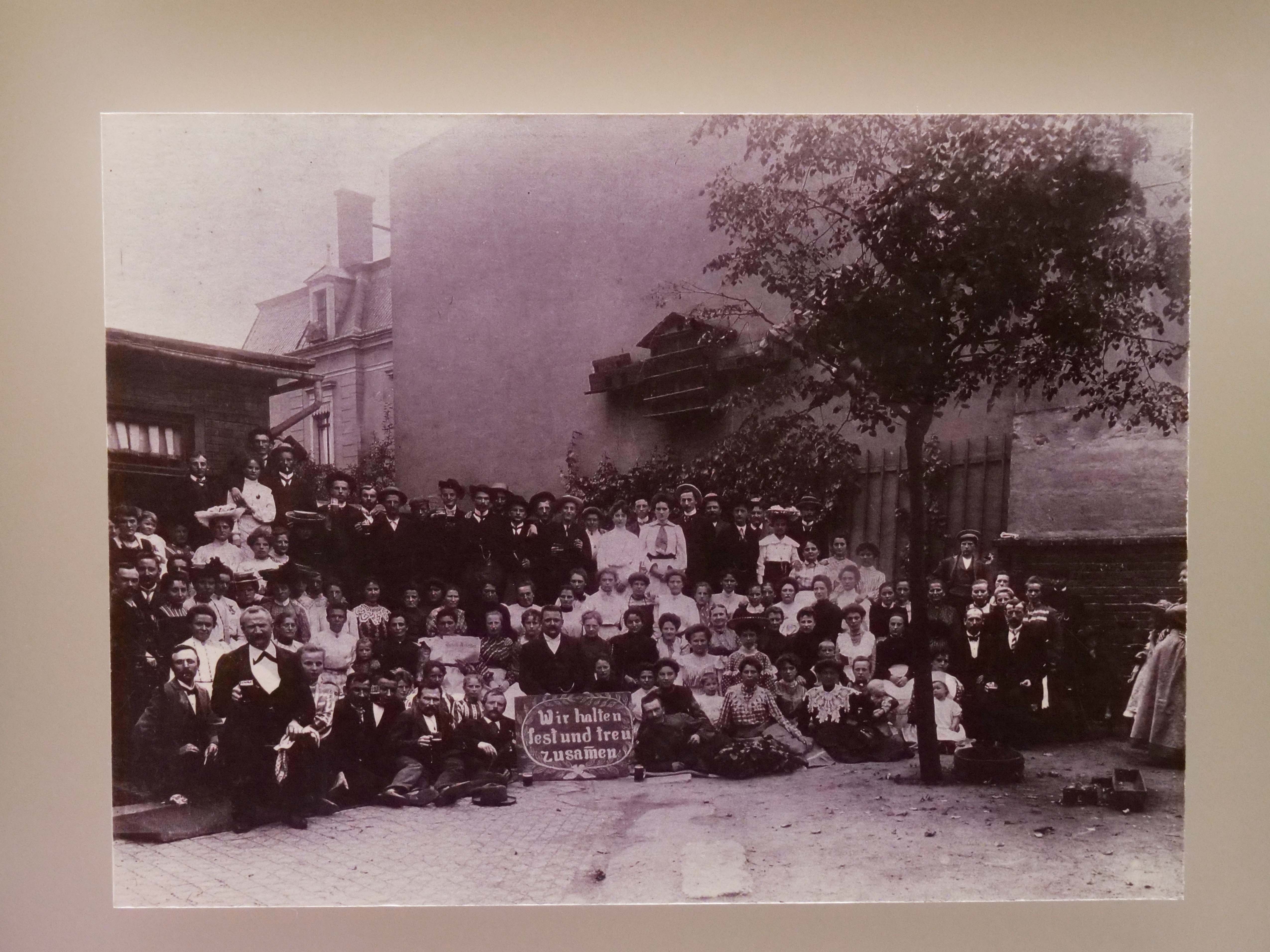 KI generiert: Das Bild zeigt eine große Gruppe von Menschen, die für ein Foto posieren, wobei einige ein Schild mit der Aufschrift "Wir halten fest und treu zusammen" halten. Die Aufnahme wirkt historisch und findet in einem städtischen Außenbereich statt.