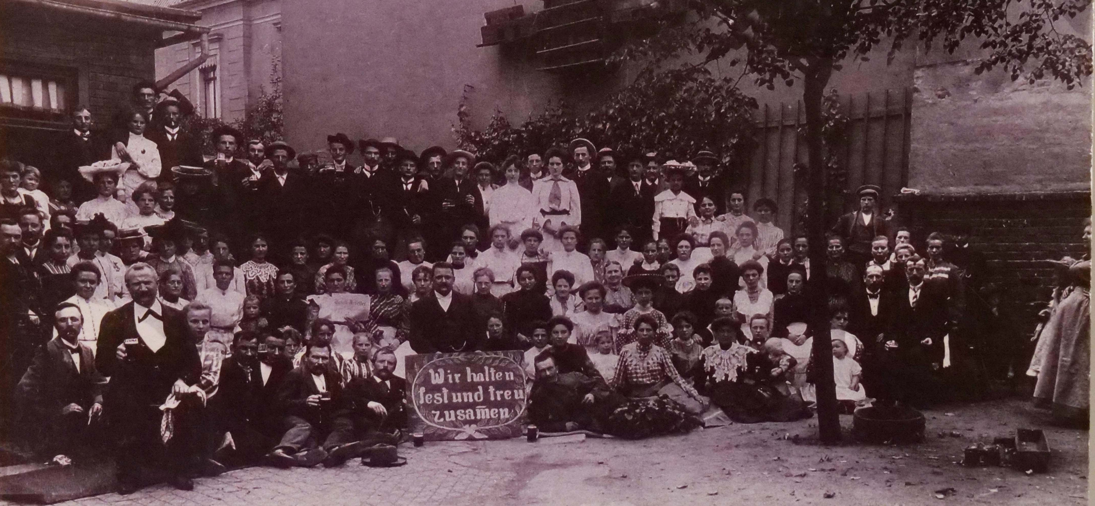 KI generiert: Das Bild zeigt eine große Gruppe von Menschen, die für ein Foto posieren, wobei einige ein Schild mit der Aufschrift "Wir halten fest und treu zusammen" halten. Die Aufnahme wirkt historisch und findet in einem städtischen Außenbereich statt.