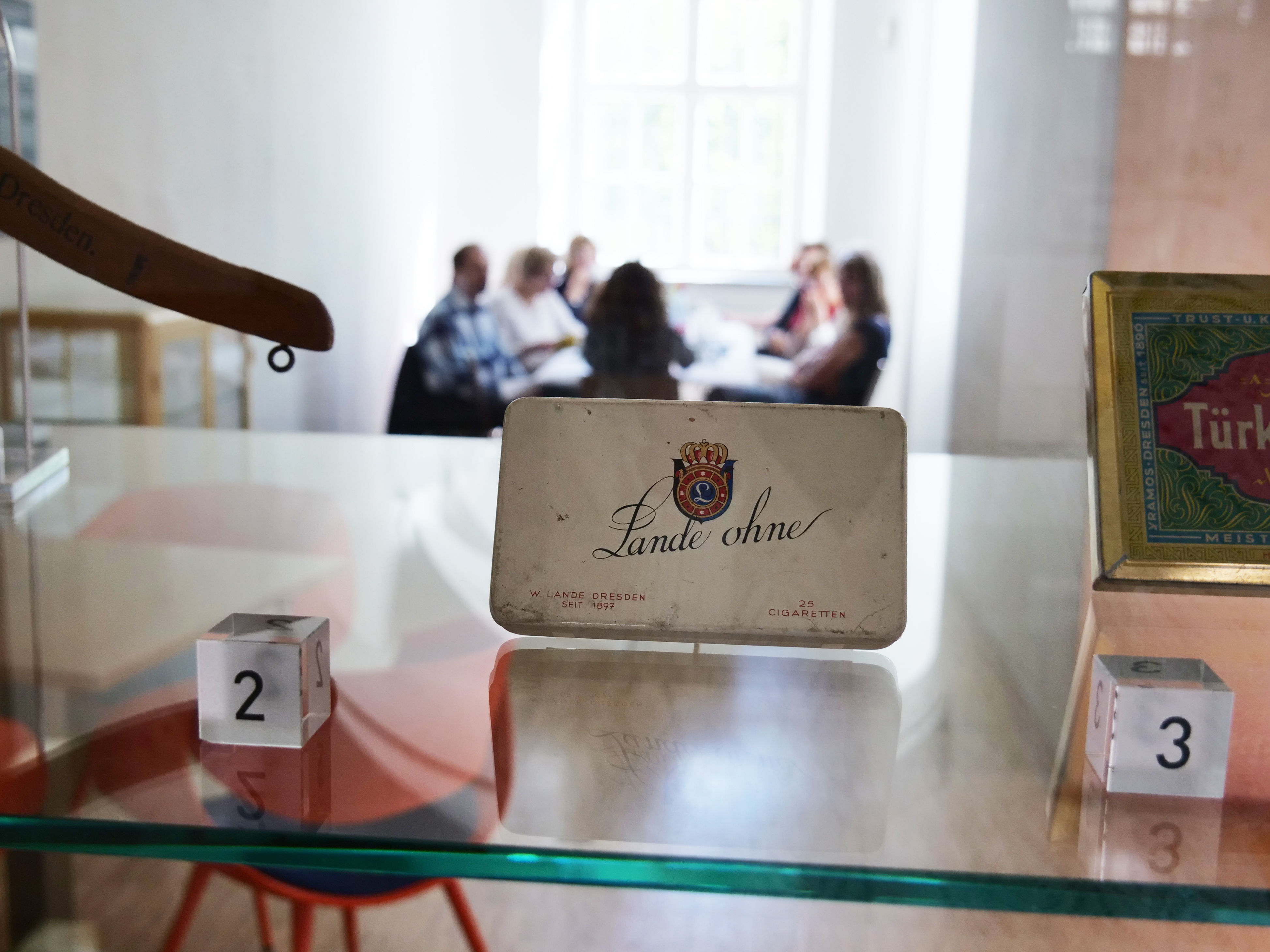 KI generiert: Das Bild zeigt eine Zigarettenschachtel in einer Vitrine, mit verschwommenen Personen im Hintergrund, die an einem Tisch sitzen. Der Fokus liegt auf der Schachtel mit der Aufschrift "Lande ohne".