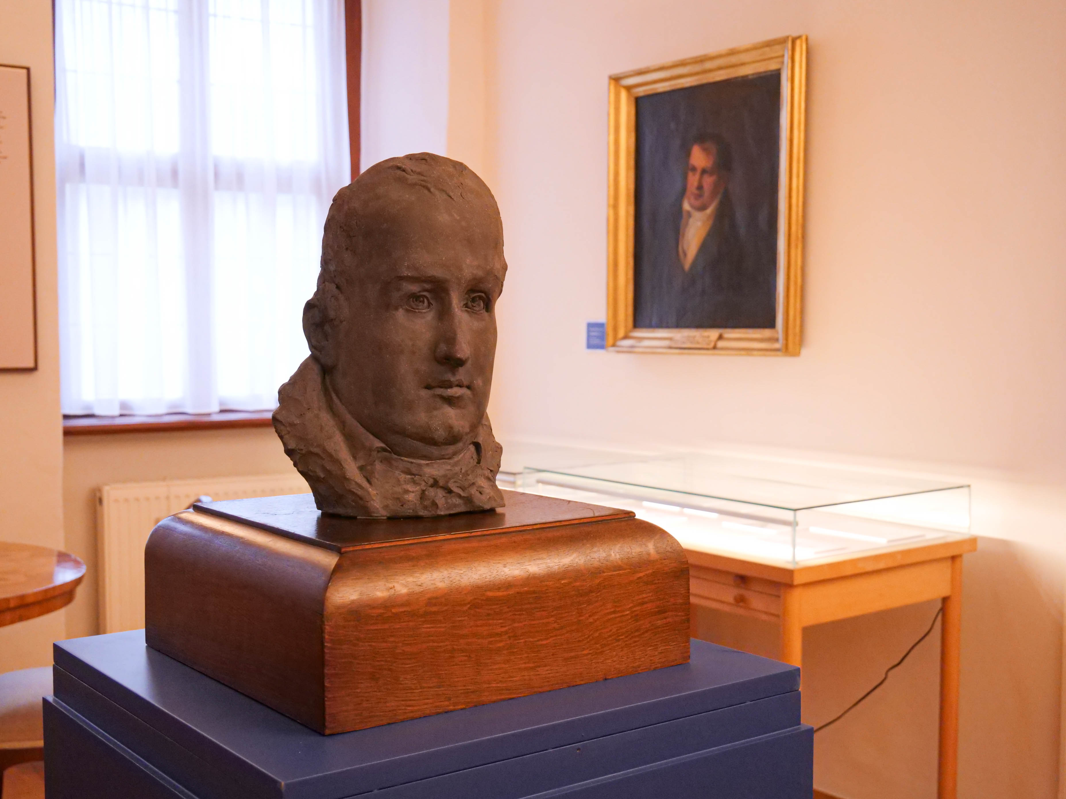KI generiert: Das Bild zeigt eine Büste eines Mannes auf einem Sockel in einem Raum, der wie ein Museum oder eine Ausstellung aussieht. Im Hintergrund hängt ein gerahmtes Porträt an der Wand.