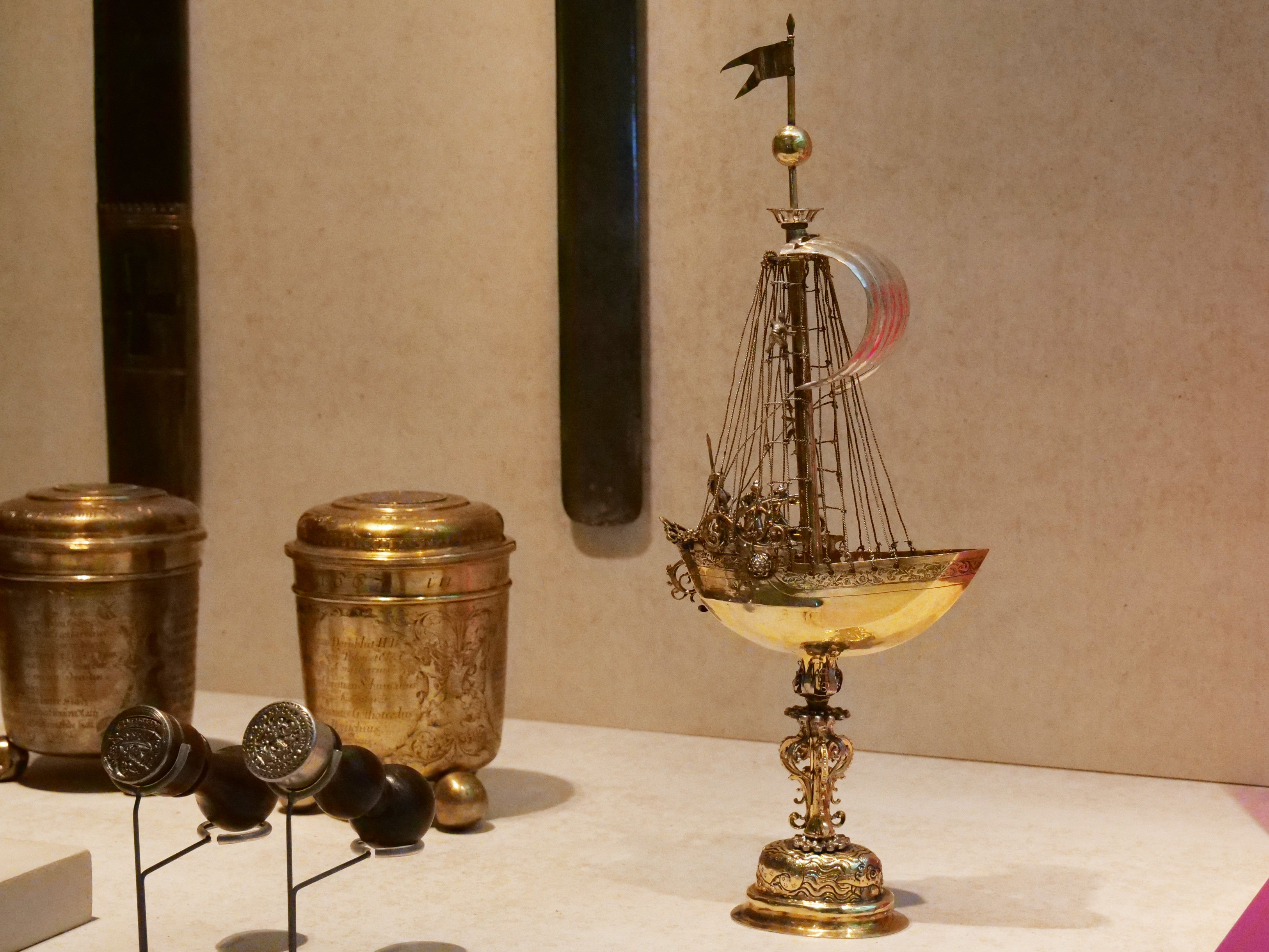 AI generated: The image shows an ornately decorated drinking vessel in the shape of a ship on a pedestal, displayed in a museum. Other antique objects can be seen around the ship.