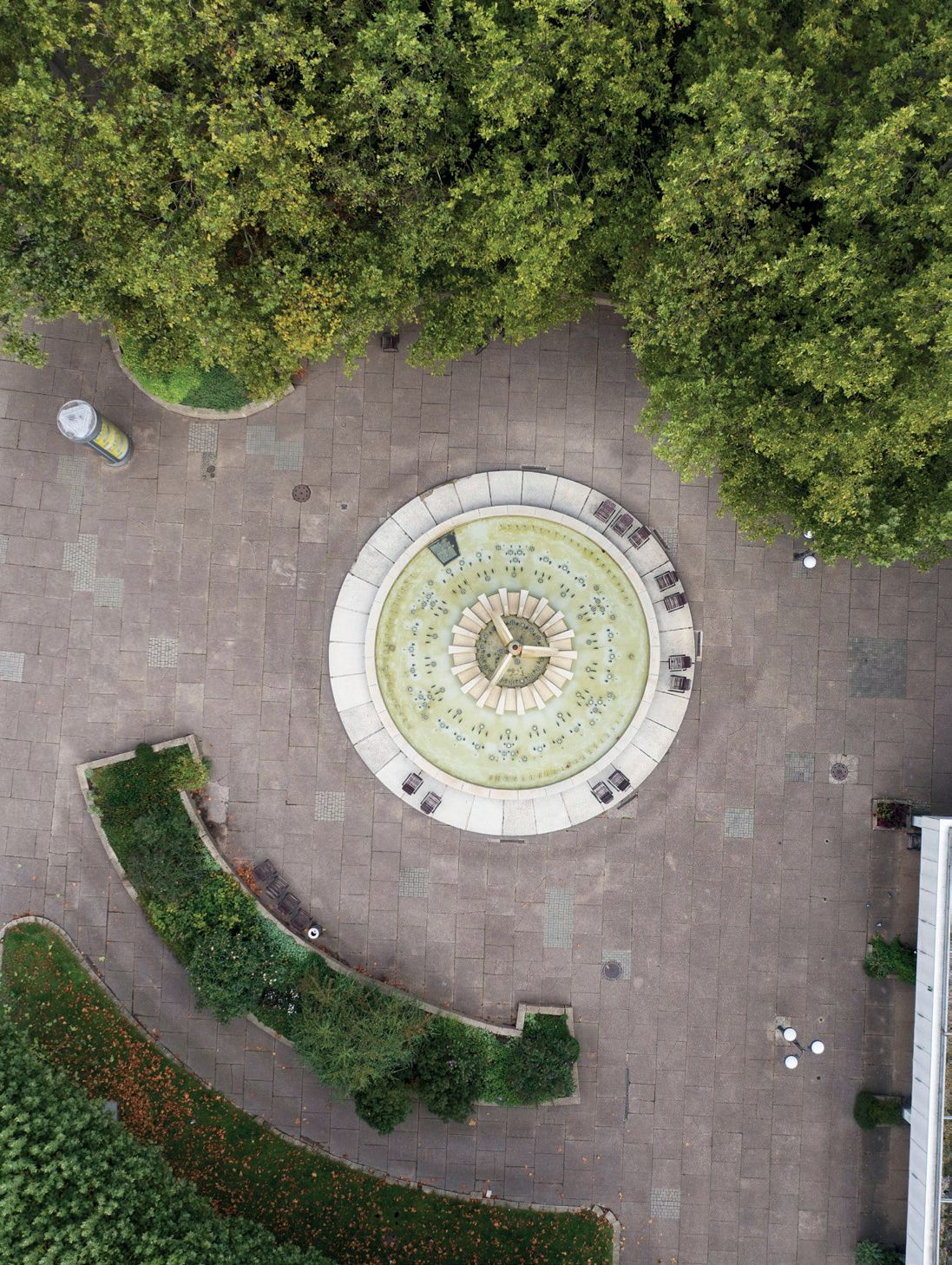 KI generiert: Das Bild zeigt eine luftaufnahme eines runden Brunnens auf einem gepflasterten Platz, umgeben von Bäumen und Grünflächen. Der Brunnen ist von oben sichtbar und steht in der Mitte des Bildes.