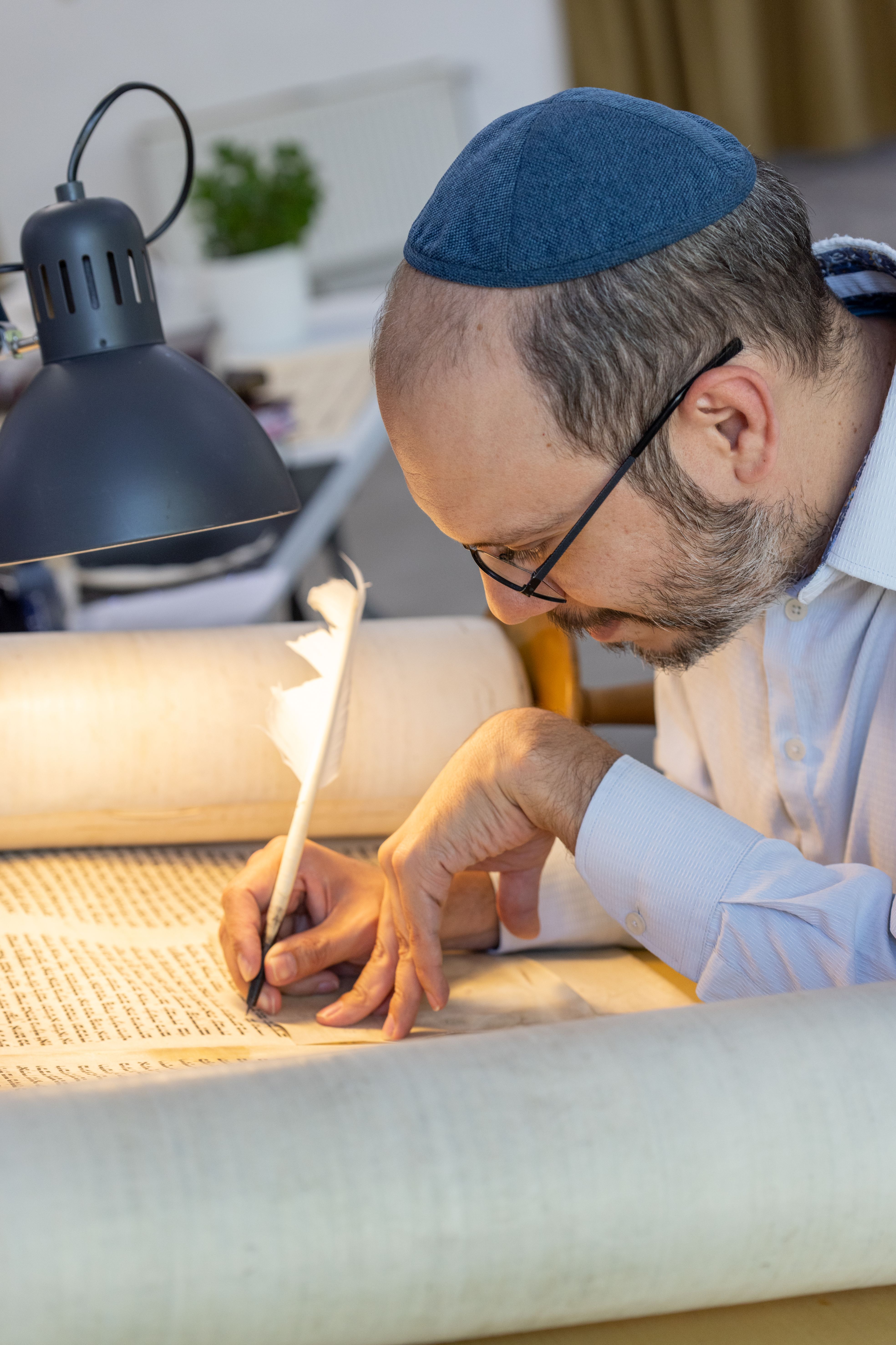 KI generiert: Das Bild zeigt einen Mann, der konzentriert mit einer Feder auf einer Schriftrolle schreibt. Er trägt eine Kippa und arbeitet unter einer Tischlampe.