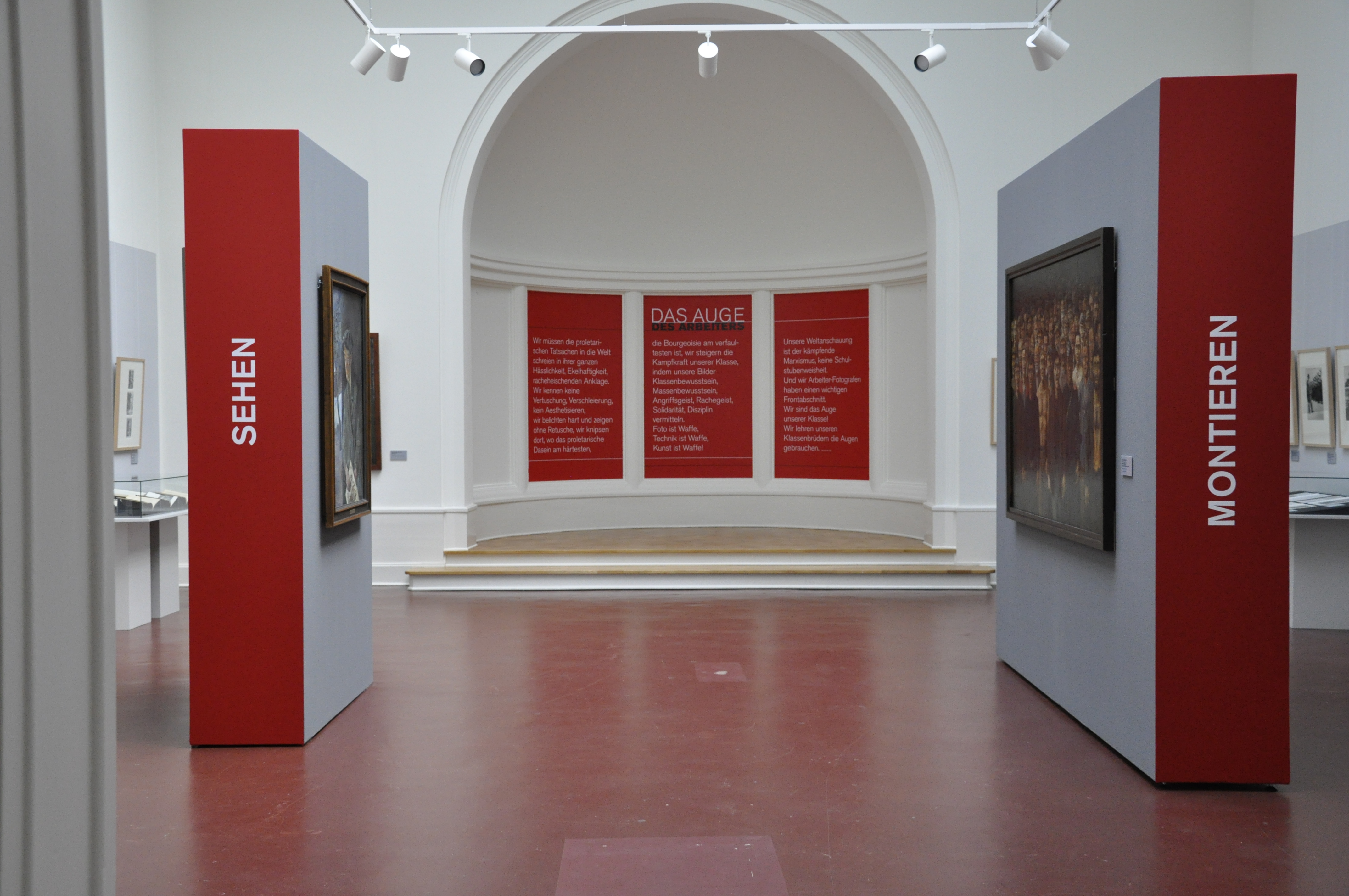 KI generiert: Das Bild zeigt eine Museumsausstellung mit Wänden, auf denen "SEHEN" und "MONTIEREN" geschrieben steht, und im Hintergrund vier rot beschriftete Tafeln unter dem Titel "DAS AUGE". Es sind Gemälde und Kunstwerke an den Wänden ausgestellt.