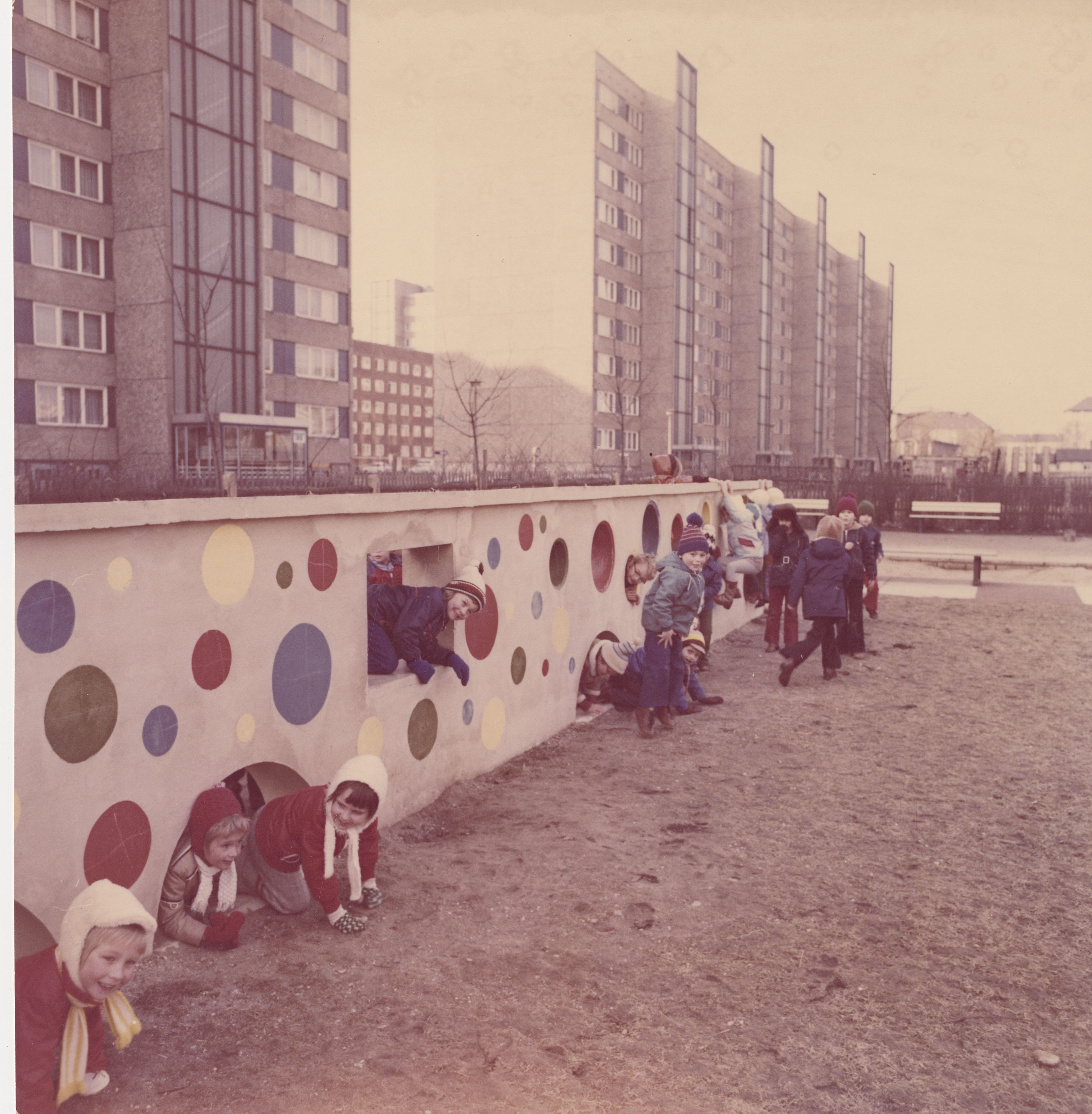 KI generiert: Das Bild zeigt eine Gruppe von Kindern, die auf einem Spielplatz spielen und durch eine bunt bemalte Mauer mit Kreisen krabbeln und klettern. Im Hintergrund sind große Wohngebäude zu sehen.