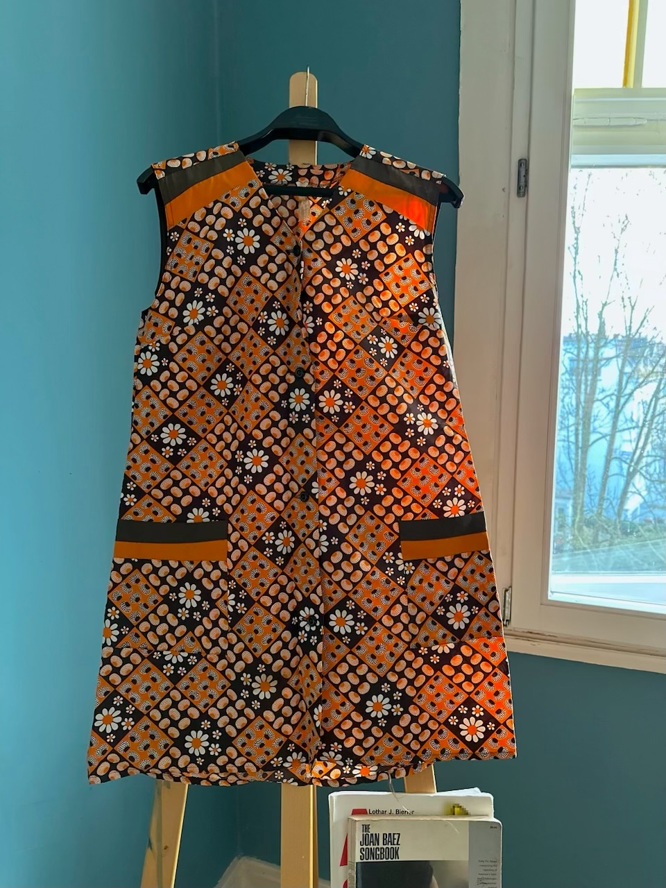 KI generiert: Das Bild zeigt ein ärmelloses, gemustertes Kleid, das an einem Kleiderbügel hängt. Der Hintergrund ist eine Wand in einem hellen Blau, neben einem Fenster.