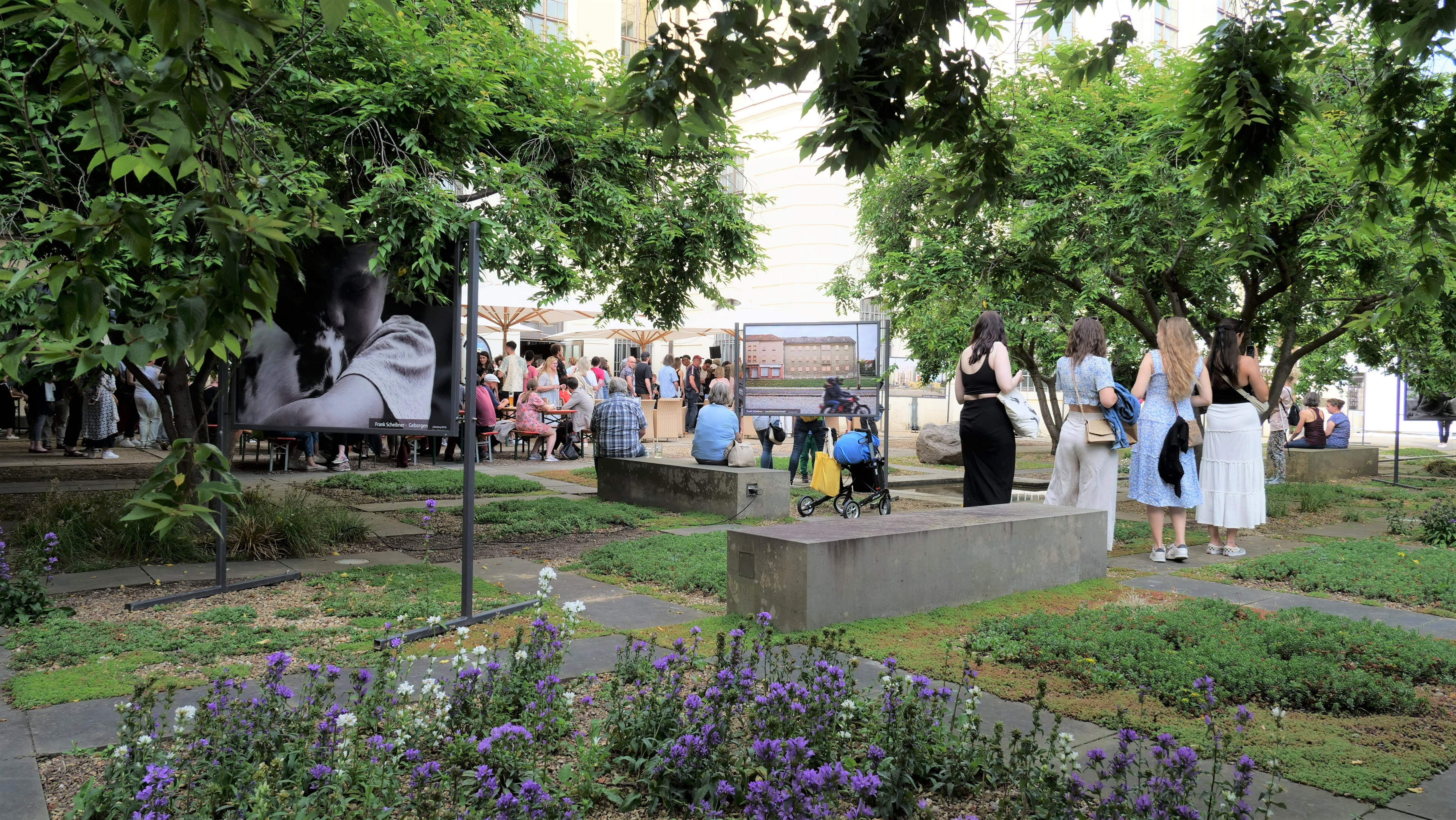 KI generiert: Das Bild zeigt eine Gruppen von Menschen, die in einem grün bepflanzten Außenbereich eine Kunstausstellung oder ein Treffen besuchen. Im Hintergrund sind Kunstwerke sowie ein Bereich mit Sitzmöglichkeiten zu sehen.