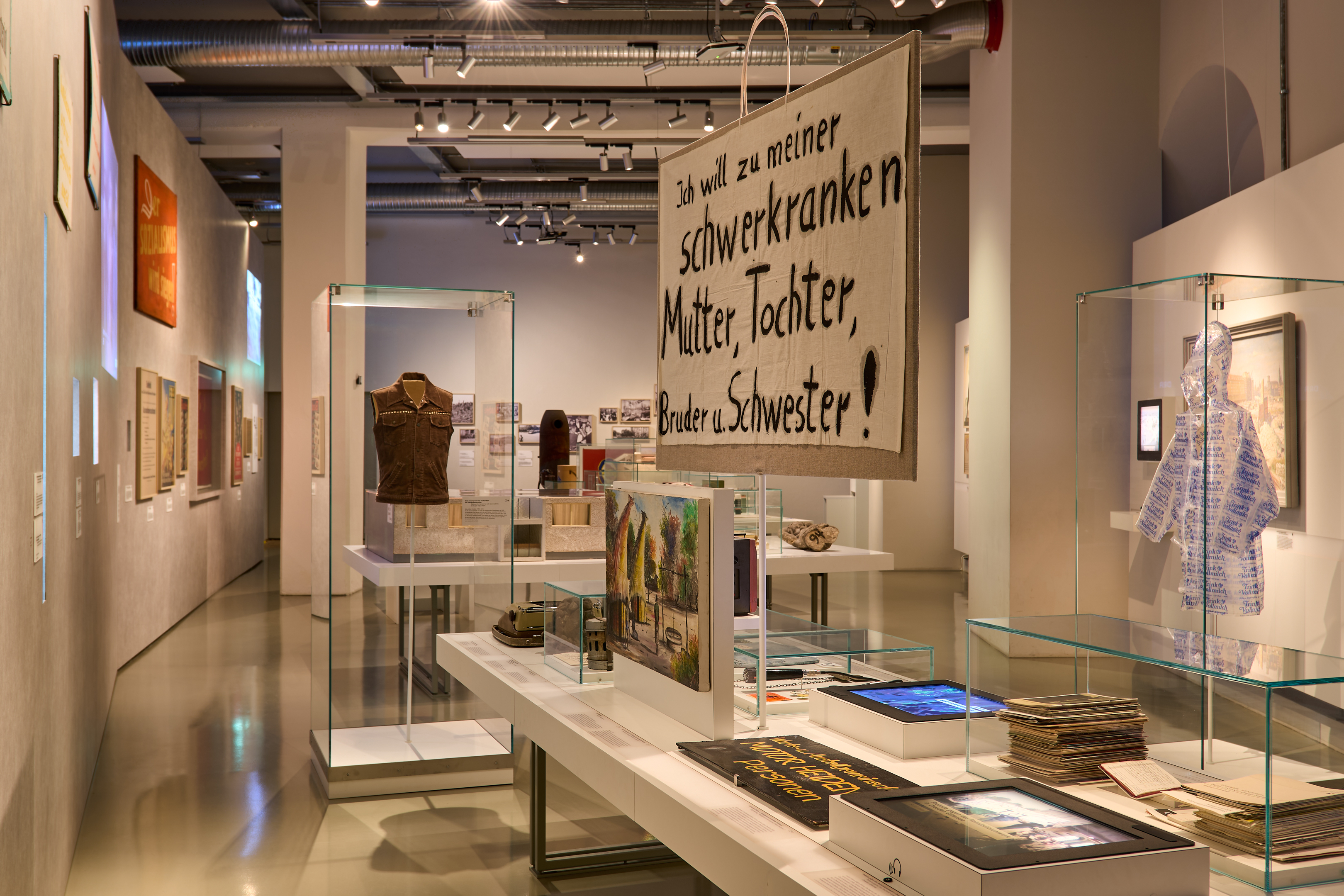 KI generiert: Das Bild zeigt eine Ausstellung in einem Museum, in der verschiedene Kunstwerke und historische Objekte präsentiert werden. Eine auffällige Installation im Vordergrund enthält ein großes Schild mit der Aufschrift "Ich will zu meiner schwerkranken Mutter, Tochter, Bruder u. Schwester!".