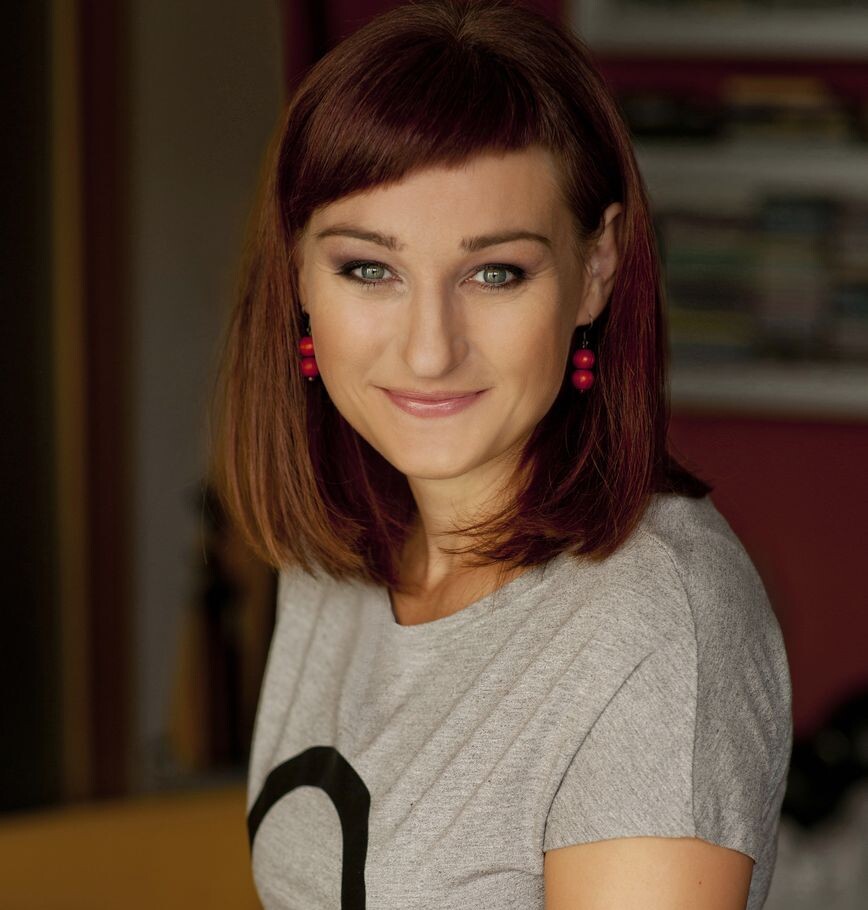 AI generated: The image shows a woman with red hair and red earrings smiling at the camera. She is wearing a grey T-shirt and is standing in an interior space.