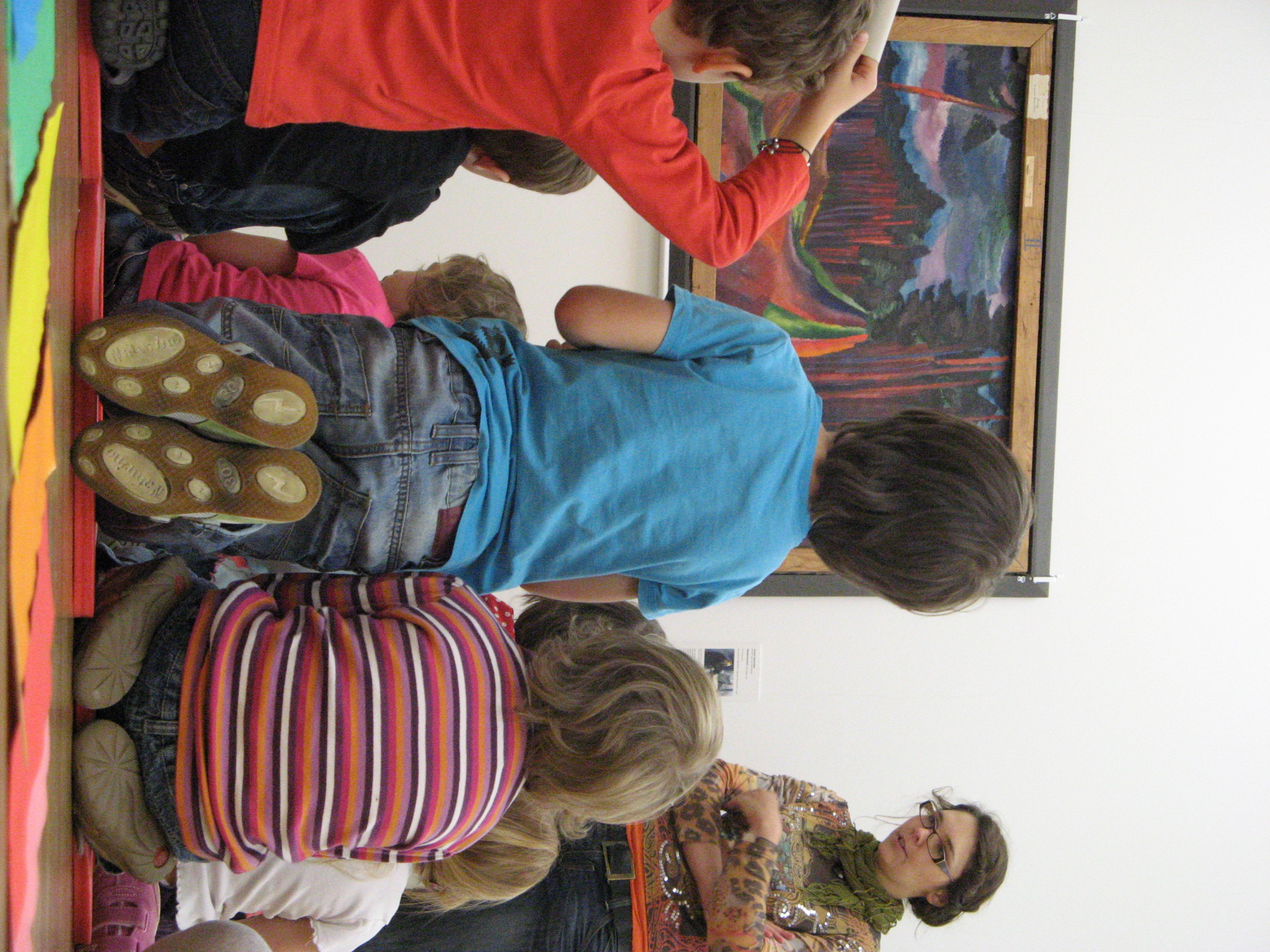 AI generated: The image shows a group of children sitting in front of a work of art in a museum or gallery and listening to or looking at it attentively. An adult is standing next to them and appears to be explaining something to them.