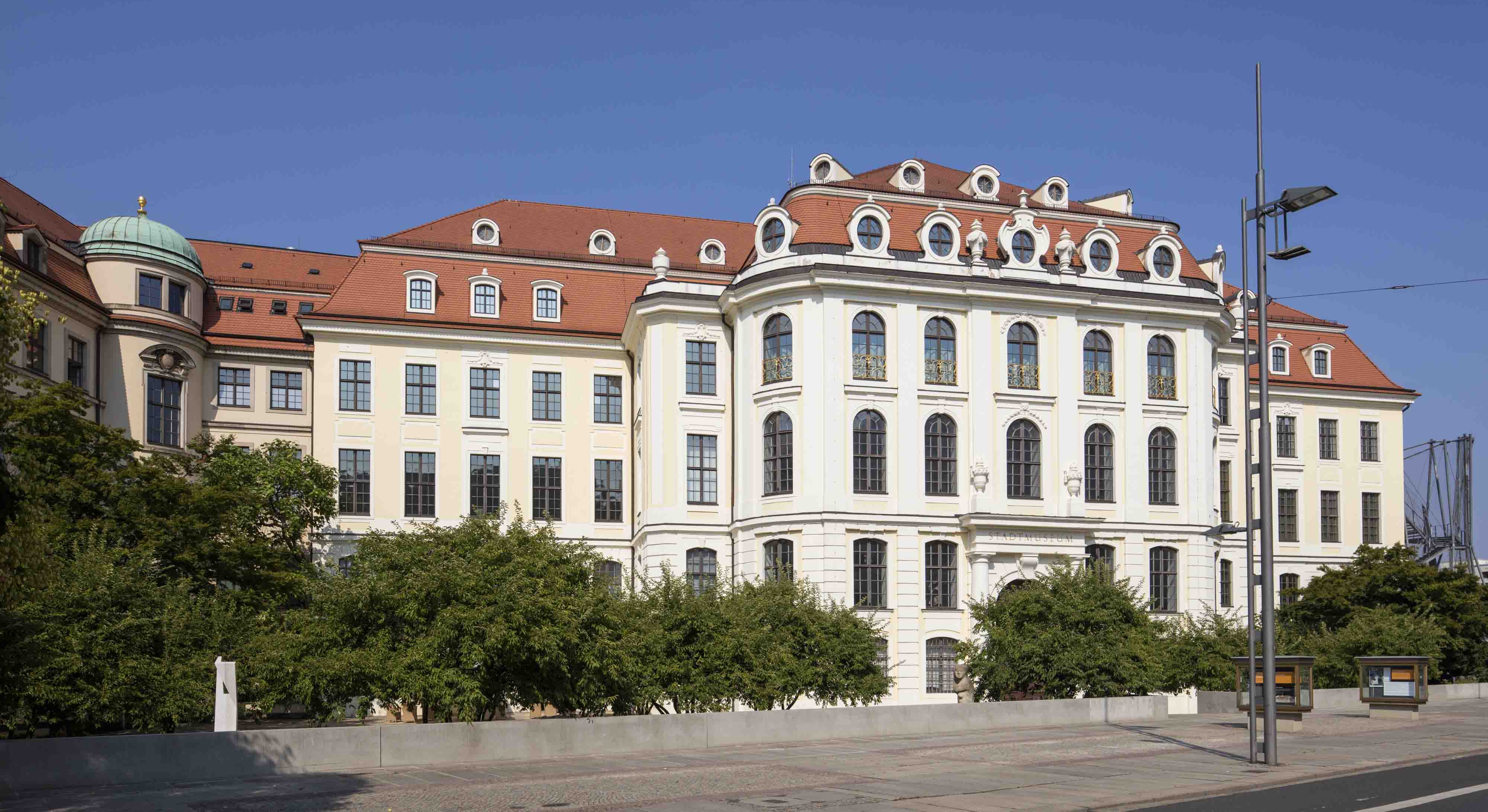 KI generiert: Das Bild zeigt ein großes, historisches Gebäude mit einem roten Dach und zahlreichen Fenstern. Vor dem Gebäude befinden sich Büsche und eine kleine Mauer.