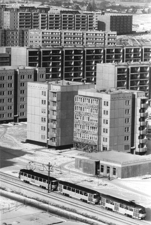 KI generiert: Das Bild zeigt eine städtische Wohngegend mit Plattenbau-Hochhäusern, die in der DDR typisch waren. Im Vordergrund fährt eine Straßenbahn an den Wohnhäusern vorbei.