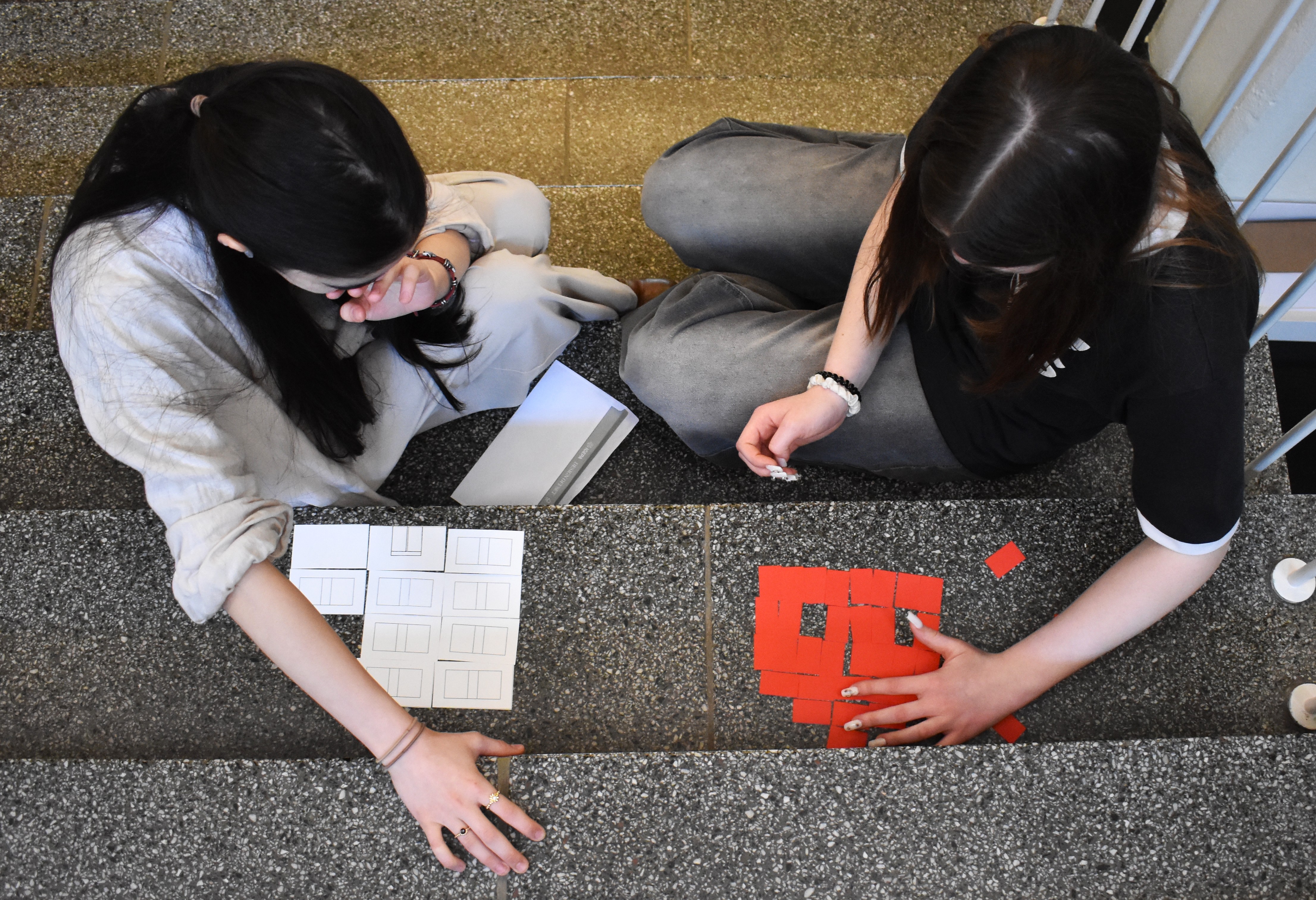 KI generiert: Das Bild zeigt zwei Personen, die auf einer Steintreppe sitzen und mit rechteckigen Papierschnipseln arbeiten. Eine Person hat weiße Schablonen vor sich, während die andere rote ausgeschnittene Stücke anordnet.