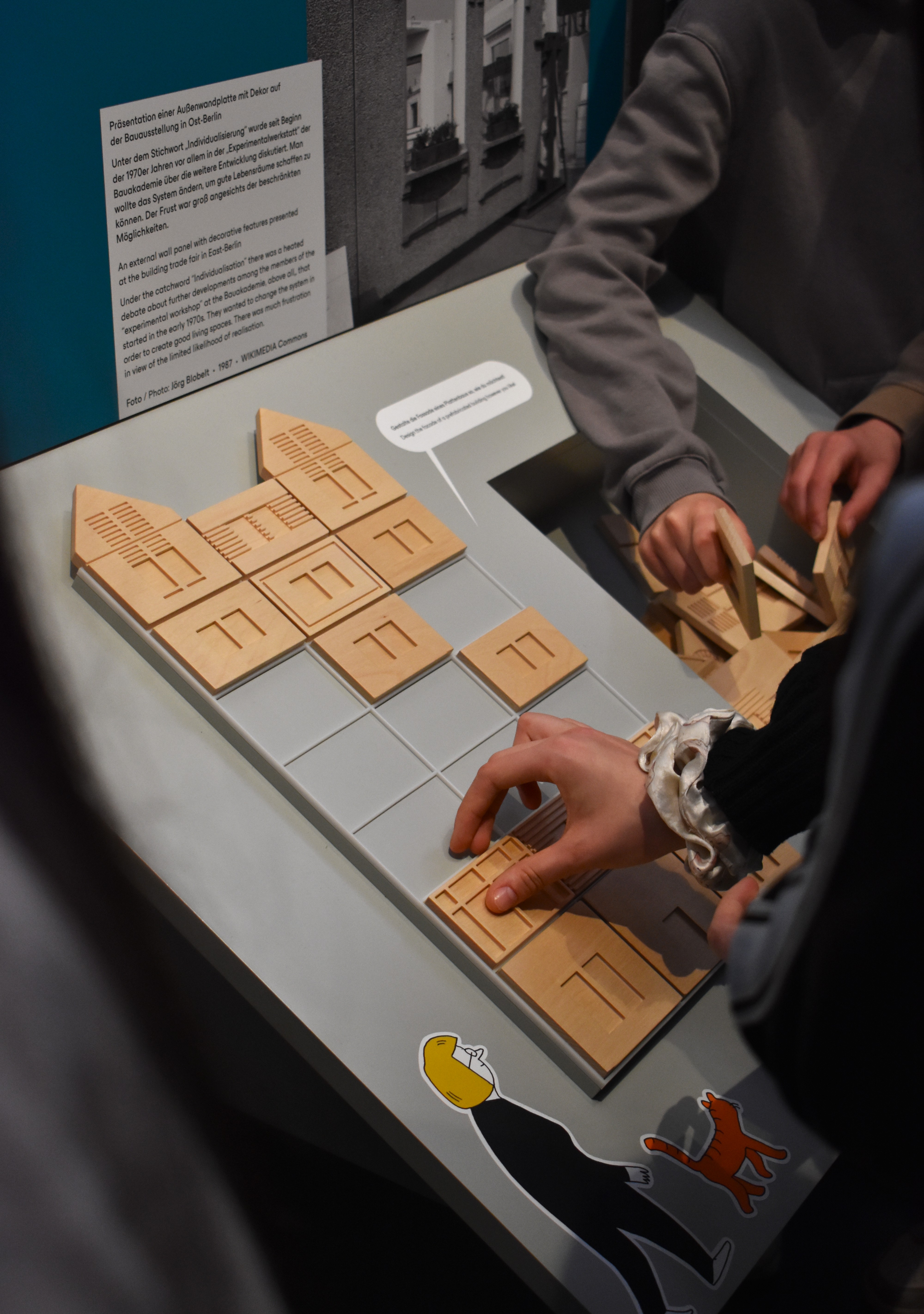 KI generiert: Das Bild zeigt ein interaktives Display, auf dem Personen Holzklötze platzieren, um möglicherweise ein Gebäude oder ein Stadtbild nachzustellen. Es scheint sich um eine Ausstellung zu handeln, die sich mit Architektur oder Stadtplanung beschäftigt.