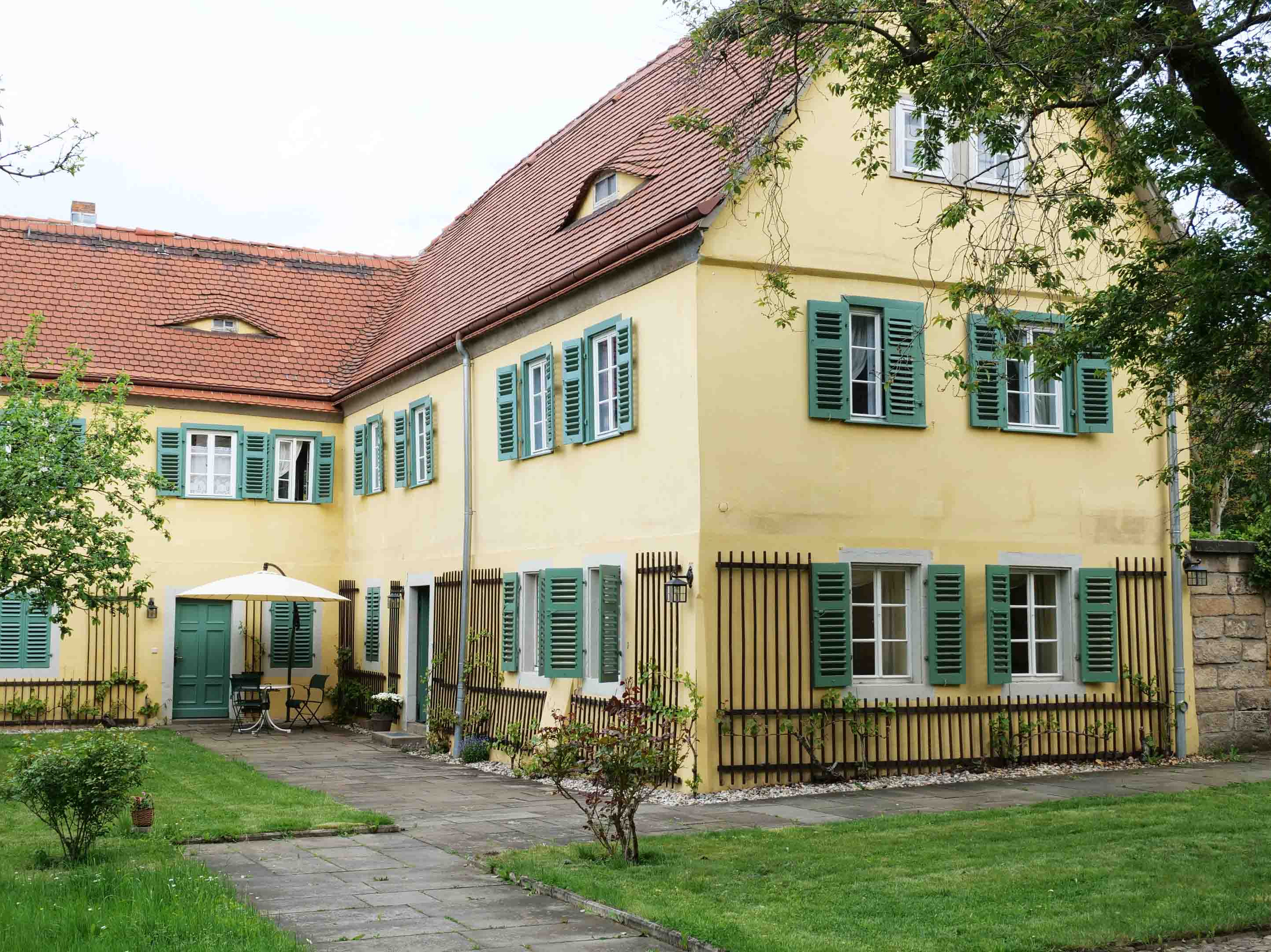 KI generiert: Das Bild zeigt ein großes, zweistöckiges, gelb gestrichenes Haus mit grünen Fensterläden und einem roten Ziegeldach, das sich in einem gepflegten Garten mit gepflastertem Gehweg befindet. Der Garten hat eine Terrasse mit einem Sonnenschirm und Gartenmöbeln unter einem Baum.