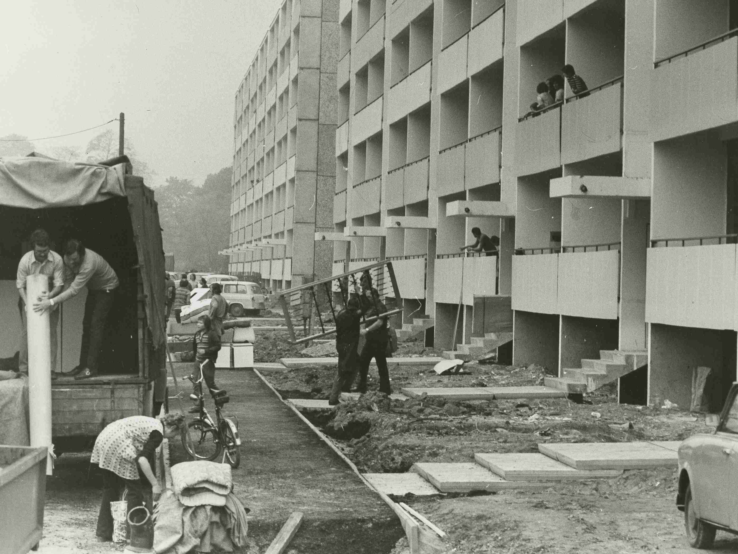 AI generated: The image shows a construction site in front of a multi-storey residential building, with several people busy with construction work and material deliveries. In the foreground are a lorry, a bicycle and a car belonging to the construction site.