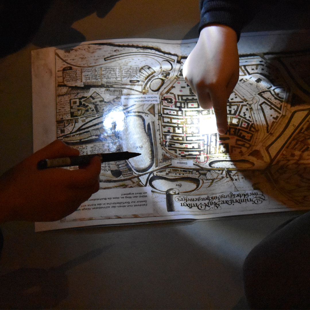 AI-generated: The image shows two hands pointing at an old town map illuminated by a beam of light. One hand is holding a pen, whilst the other is pointing to a specific spot on the map.