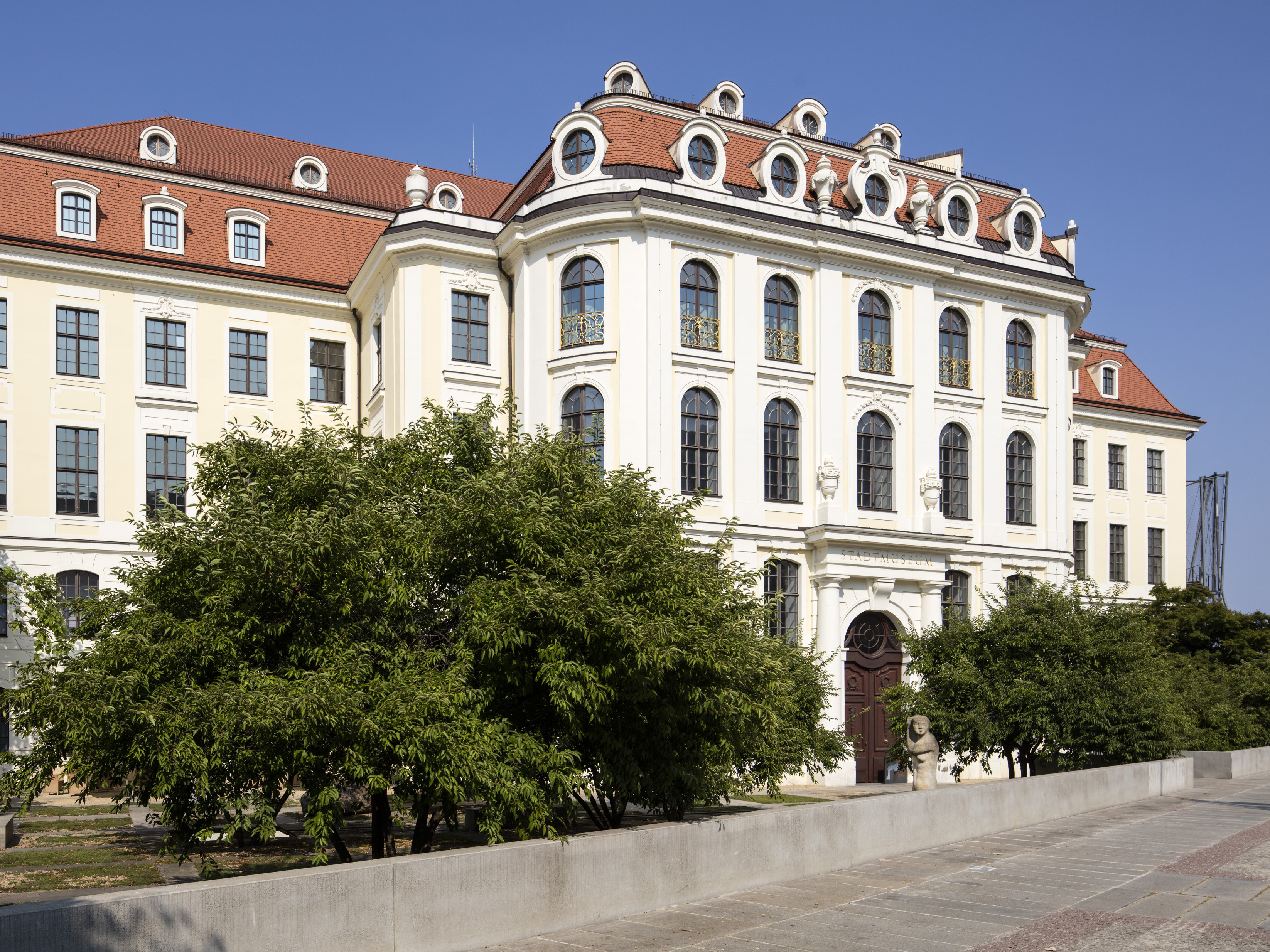 KI generiert: Das Bild zeigt ein imposantes historisches Gebäude mit einer reich verzierten Fassade und zahlreichen großen Fenstern. Vor dem Gebäude sind mehrere Bäume und Sträucher zu sehen.