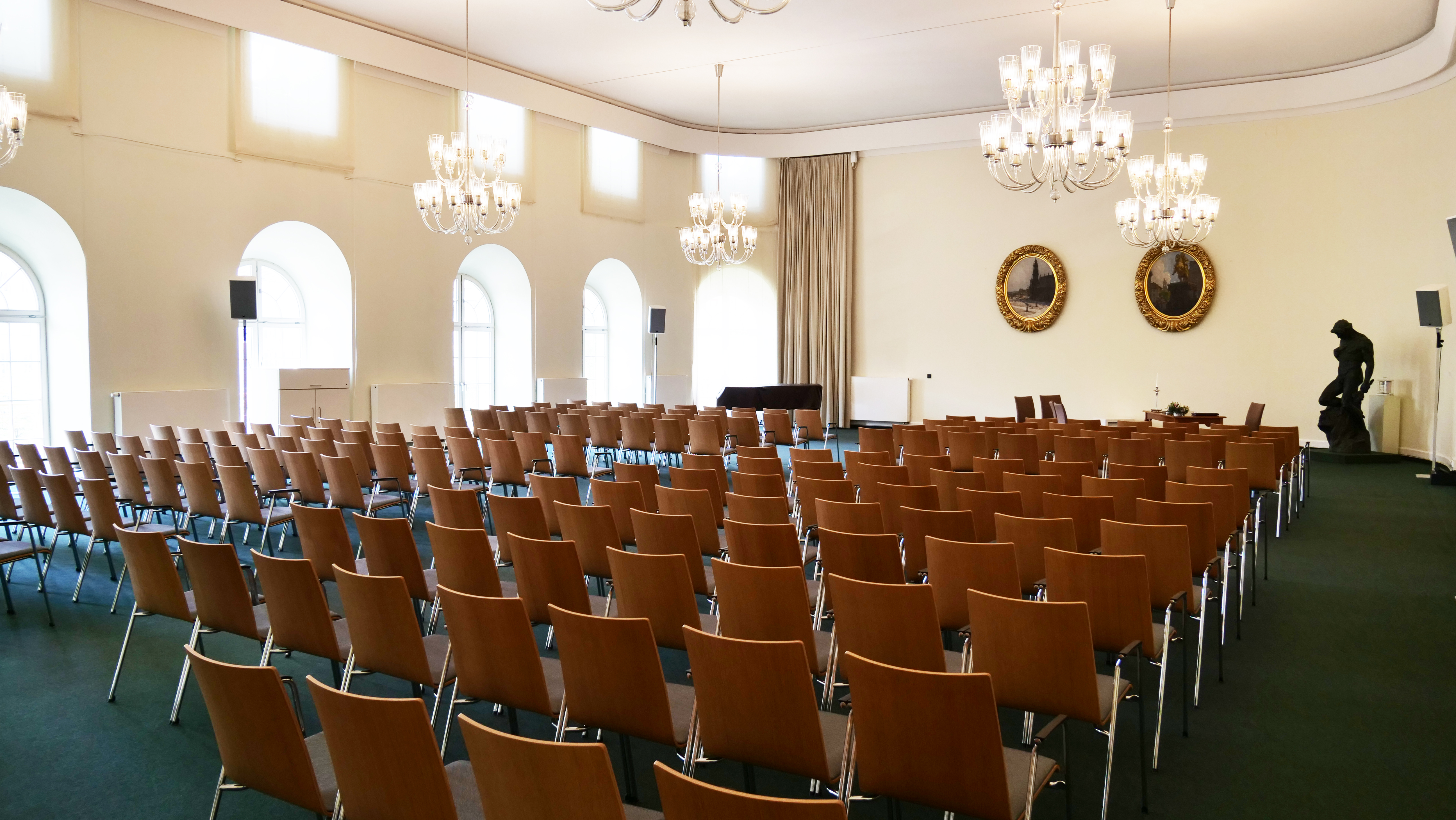 KI generiert: Das Bild zeigt einen großen Raum, der für eine Versammlung oder ein Meeting vorbereitet ist, mit vielen Holzstühlen, die in Reihen aufgestellt sind. Der Raum ist elegant dekoriert mit Kronleuchtern, großen Fenstern und zwei Bildern an der Wand.