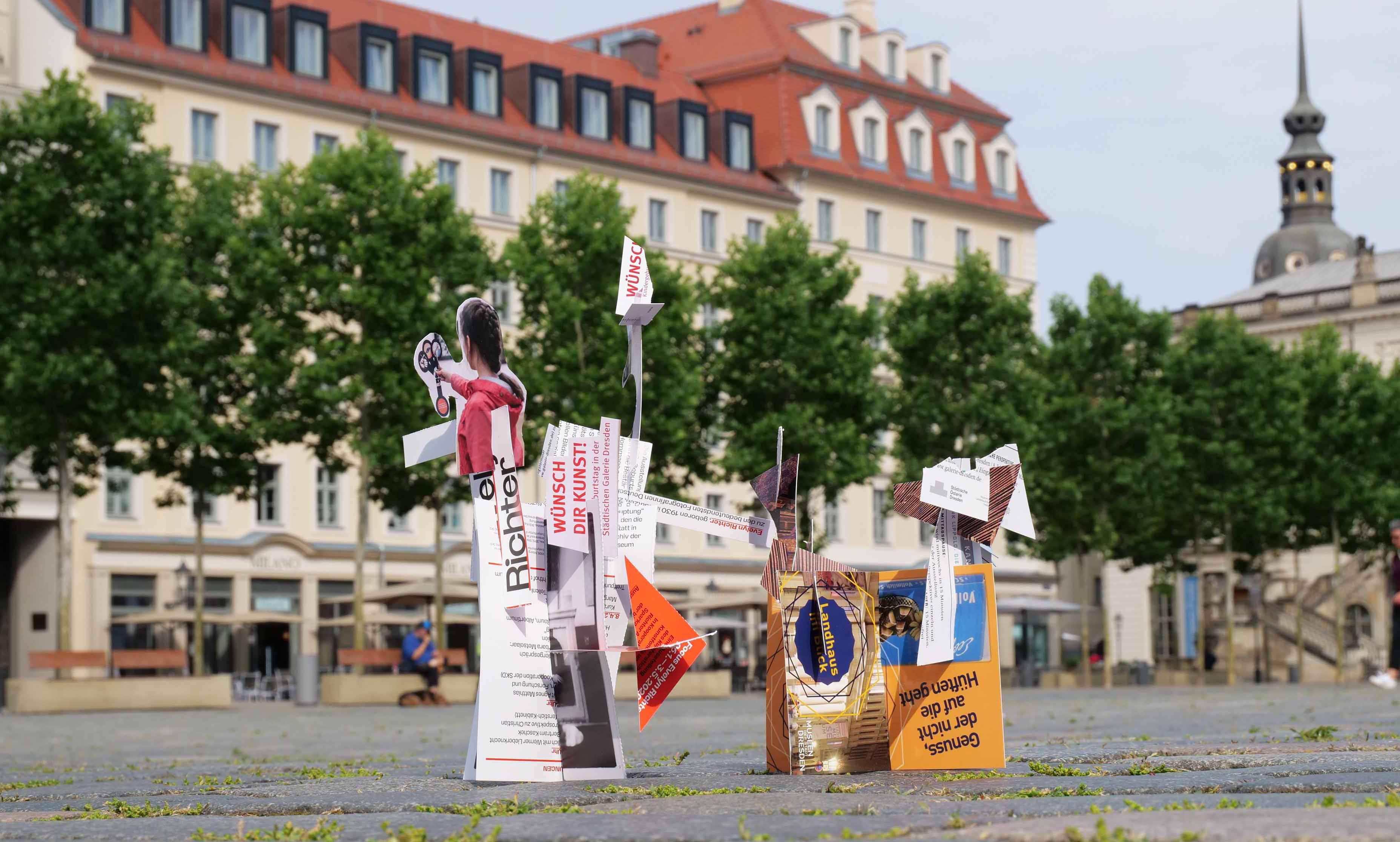 KI generiert: Das Bild zeigt eine Installation auf einem Platz, bestehend aus einer Vielzahl von bunten, papierähnlichen Objekten und Schriften. Im Hintergrund sind Gebäude und Bäume zu sehen.