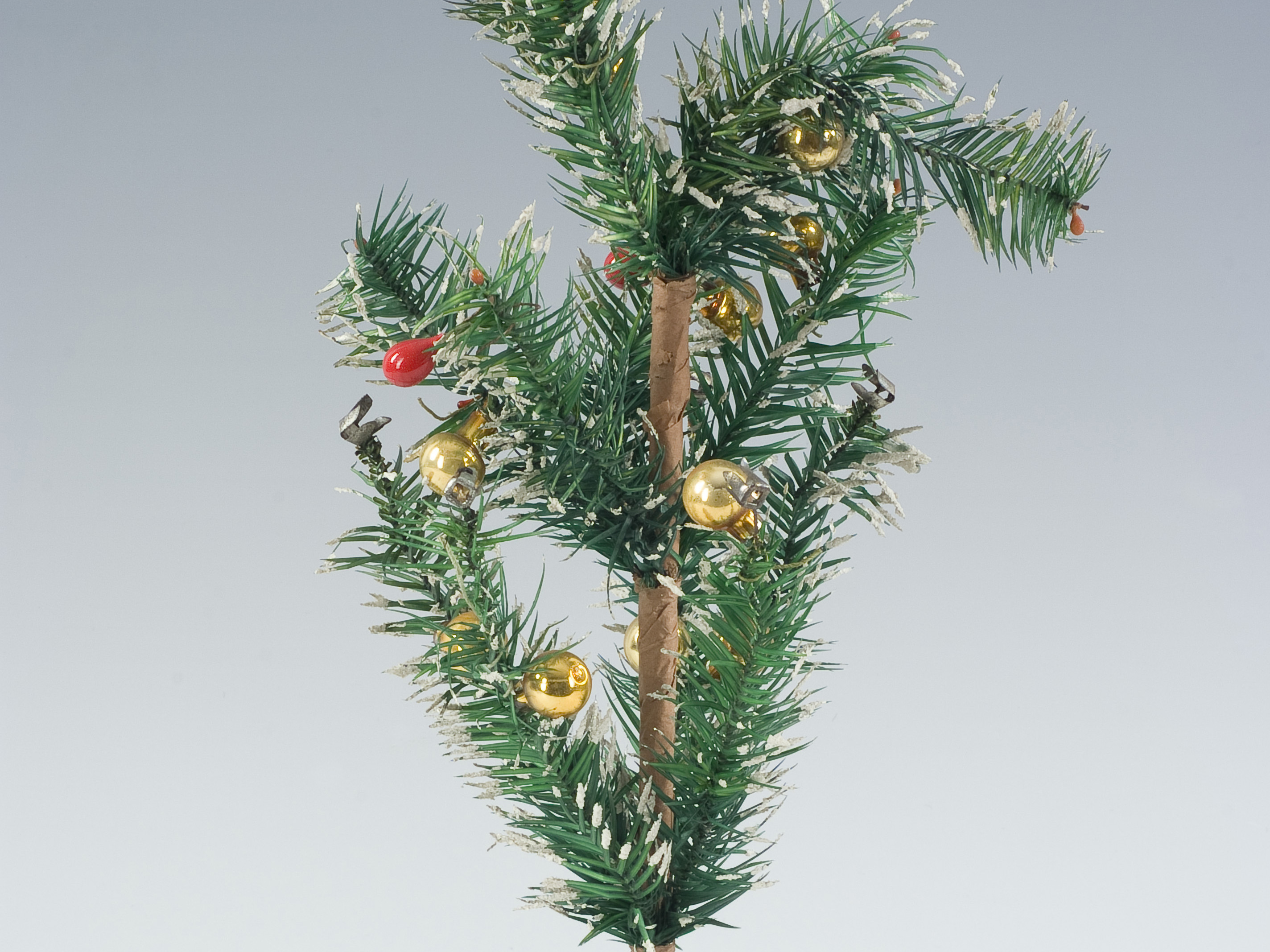 KI generiert: Das Bild zeigt einen kleinen künstlichen Weihnachtsbaum, der in einem einfachen Sockel steht. Der Baum ist mit einigen goldenen und roten Kugeln geschmückt.