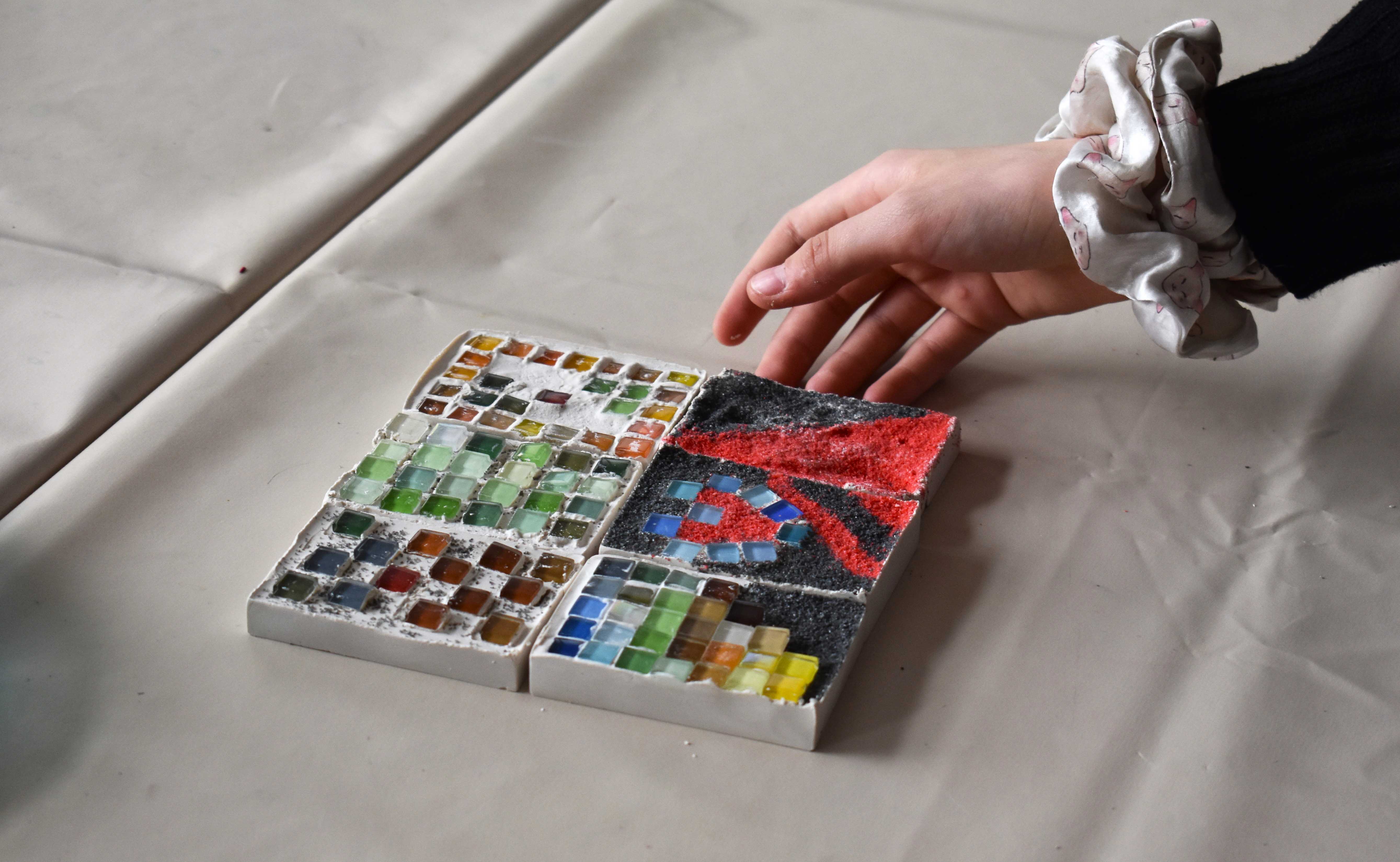 AI generated: The image shows a hand approaching a mosaic of coloured glass stones and sand. The mosaic consists of four squares with different patterns and colours.