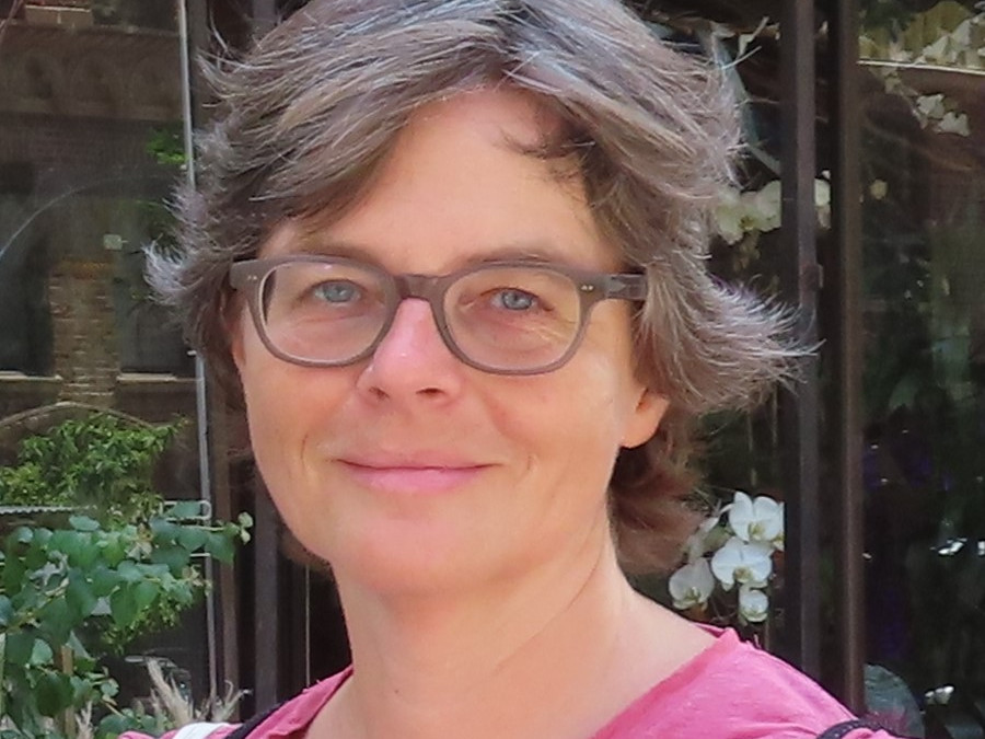 KI generiert: Das Bild zeigt eine Person mit kurzen Haaren und Brille, die ein rosa Oberteil trägt. Im Hintergrund sind Pflanzen und eine Glasfront zu sehen.