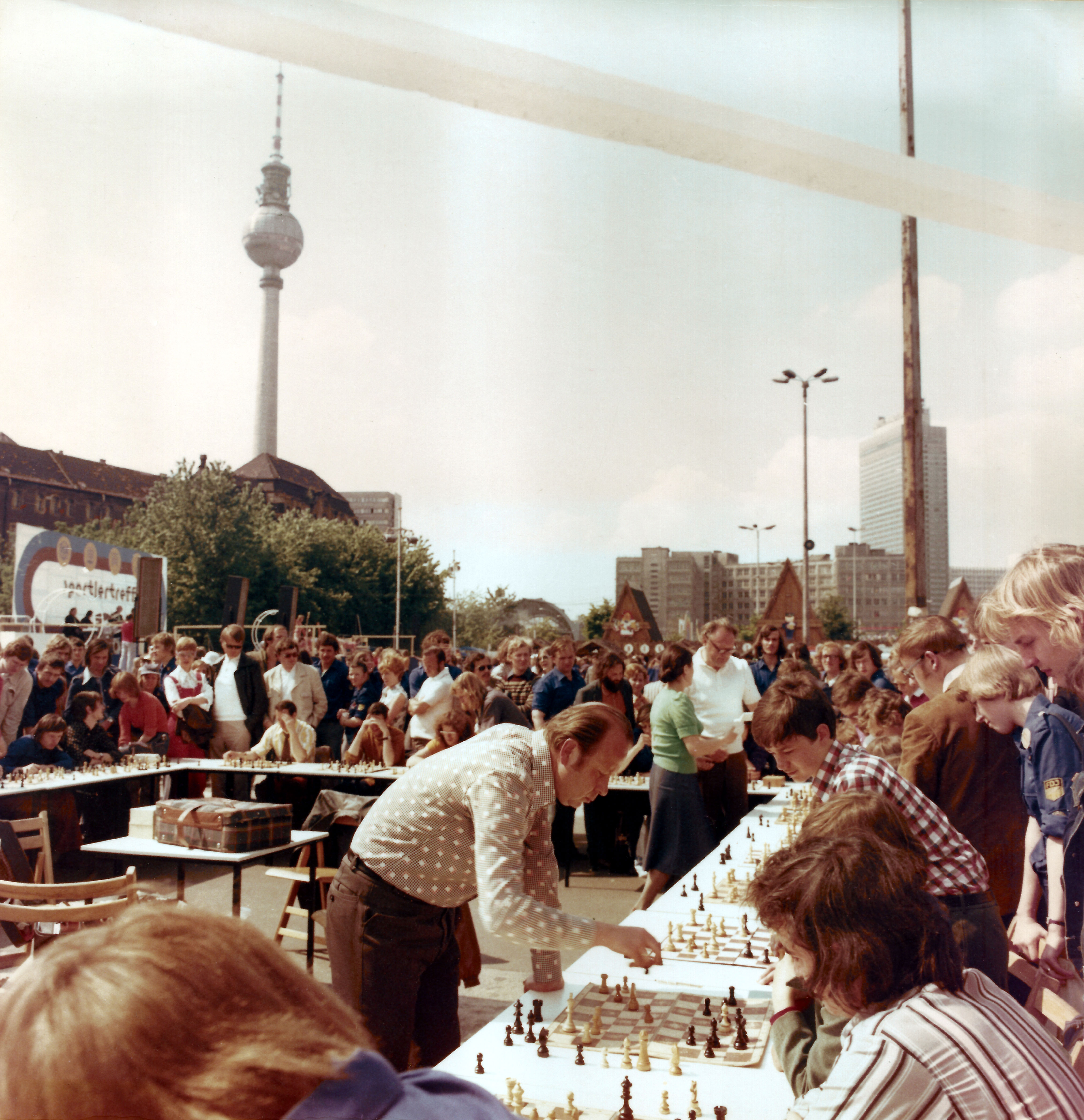 KI generiert: Das Bild zeigt ein Freiluft-Schachspiel mit vielen Zuschauern, bei dem ein Spieler mehrere Gegner an verschiedenen Brettern gleichzeitig herausfordert. Im Hintergrund ist ein Fernsehturm sichtbar, was auf einen urbanen Schauplatz hinweist.