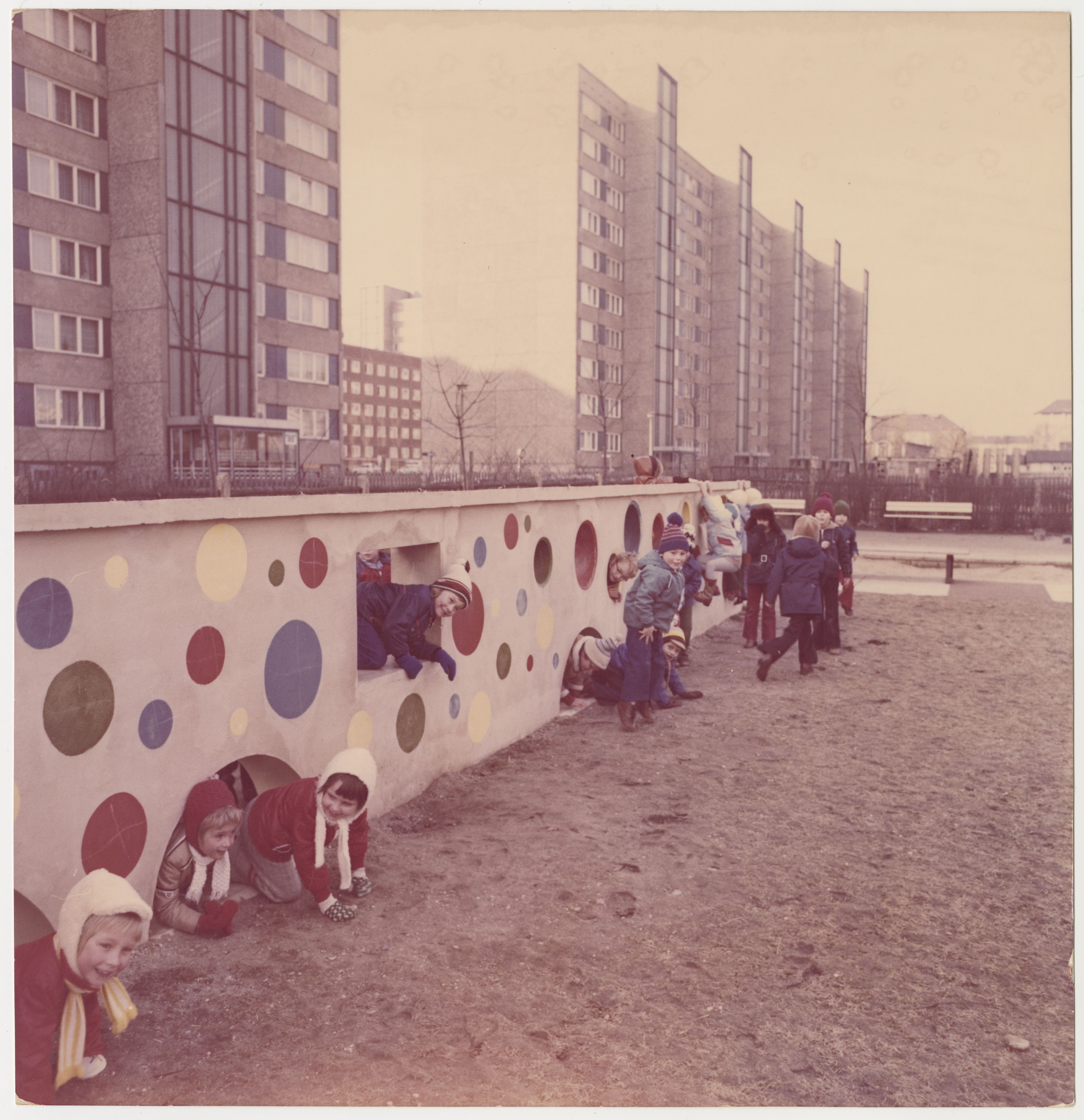 KI generiert: Das Bild zeigt eine Gruppe von Kindern, die auf einem Spielplatz spielen und durch eine bunt bemalte Mauer mit Kreisen krabbeln und klettern. Im Hintergrund sind große Wohngebäude zu sehen.